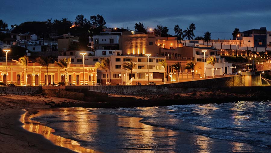 Panoramic view of a sandy beach with a variety of beachfront properties visible along the waterfront, including houses, condos, and hotels in different sizes, styles, and architectural designs. The sun is setting, casting a warm, golden glow over the scene